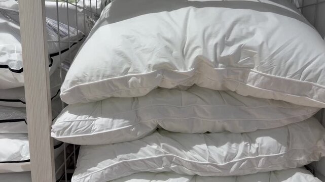 Multiple soft white pillows and folded duvets neatly stacked on metal shelves for sale in a department store, showcasing a wide selection of bedding options for home comfort and decor