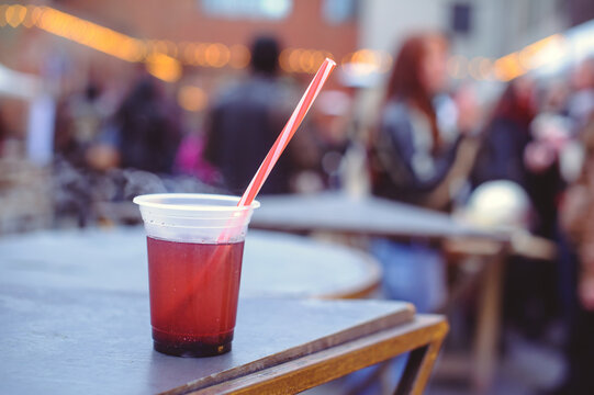 A horizontal shot of a plastic cup filled with steaming red mulled wine and a red straw on a wooden table. The background is a blurred outdoor street market with people and festive lights.