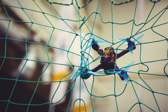 A horizontal shot of a small FPV racing drone with blue propellers stuck in a green safety net during an indoor flight.