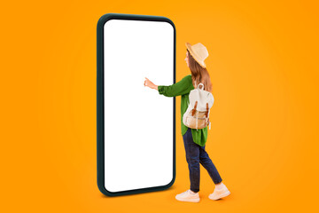 A person stands next to a tall smartphone on an orange background. She touches the screen with one hand and wears a hat and backpack. Sunlight brightens the scene. © Prostock-studio