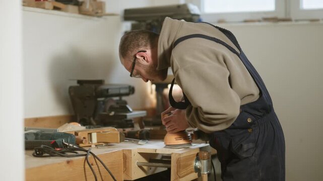 A focused man is sanding a piece of wood with an orbital sander in a workshop. He is carefully working on the surface of the wooden material.
