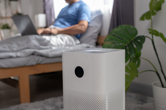Modern air purifier in bedroom with senior man resting and using laptop, clean air technology, home healthcare concept, reducing dust, allergy and pollution for healthy indoor living