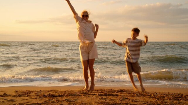 Active family jumping and dancing with joy on beach during sunset. Mom and boy celebrate on sand with raised hands. Expression of happiness success vitality excitement.