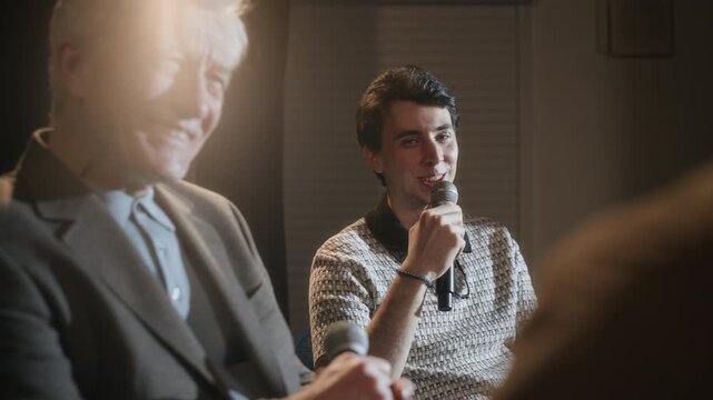 Rack focus of senior male comedian reacting on joke during comedy panel show conversation with younger colleague laughing along on stage under spotlight