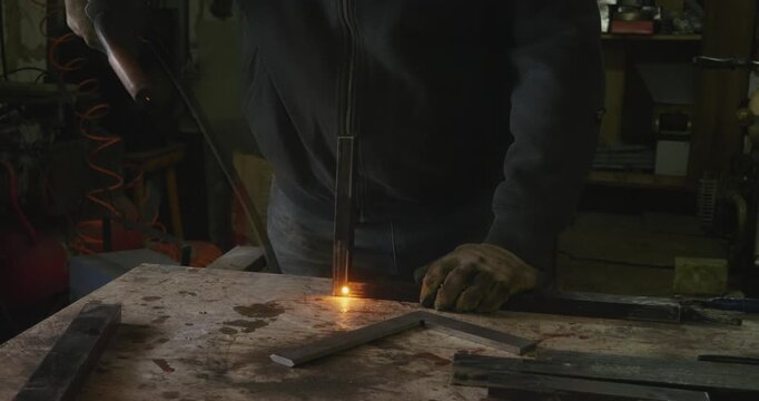Industrial welder with protective gloves using a welding machine to join metal parts in a workshop, creating bright sparks and smoke during the manufacturing process of a steel structure.