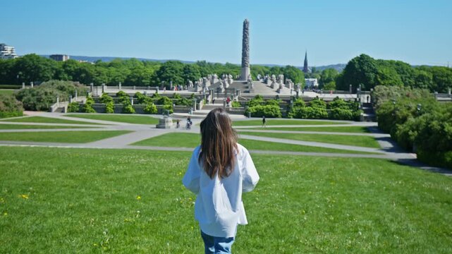 Tourist woman walking across green lawn toward the Monolith sculpture in Vigeland Sculpture Park. Monolith tower, sculpture garden and park landscape during summer in Oslo, Norway.