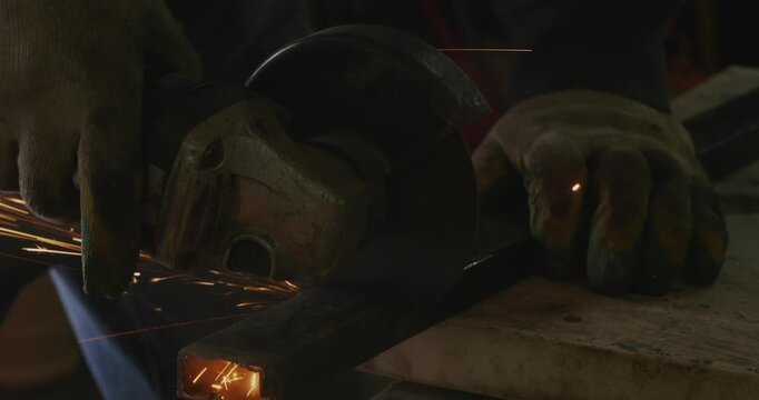 Close-up of a metalworker's hands in protective gloves using an angle grinder to cut a square metal pipe, creating a shower of bright sparks in the workshop.