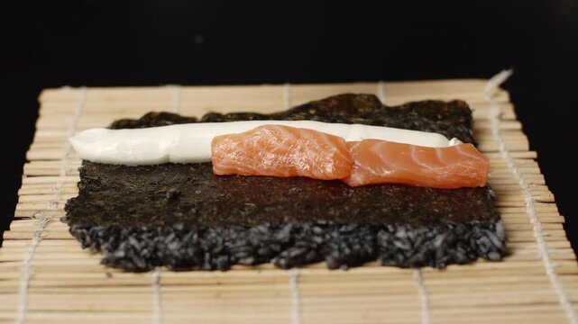 Sushi chef preparing black rice uramaki with salmon and philadelphia cream cheese on a bamboo mat, showcasing traditional japanese seafood cuisine preparation process