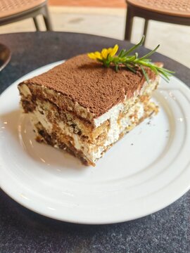 A tempting slice of tiramisu cake on a white plate, topped with cocoa powder and a delicate flower. Perfect for any coffee break or special occasion.