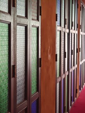 A perspective view along a row of traditional Thai-style folding windows with multi-colored glass panes and rich wooden frames.