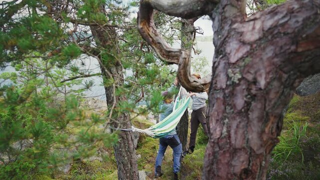 Couple setting up a hammock between pine trees on a rocky sea coast. Authentic outdoor lifestyle, forest bathing and mental wellness concept in a natural Scandinavian landscape.