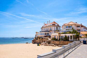 Obraz premium Beachfront villas on rocky shoreline above sandy beach Praia da Rainha in Cascais, Portugal. Wide angle view with clear sky, calm ocean, and Mediterranean architecture in bright daylight