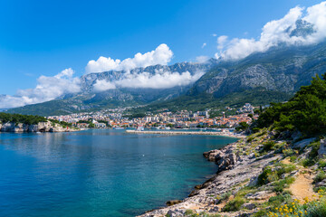 Obraz premium Scenic Mediterranean bay with coastal town, rocky shoreline and dramatic mountains under low clouds in bright summer sunlight.