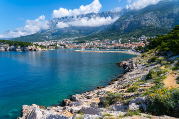Obraz premium Scenic Mediterranean bay with coastal town, rocky shoreline and dramatic mountains under low clouds in bright summer sunlight.