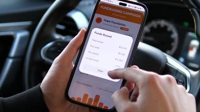 Close-up of male hands holding a smartphone inside a car. The screen displays donation statistics and a rising graph. A focused moment of reviewing fundraising progress while on the move. Ideal for