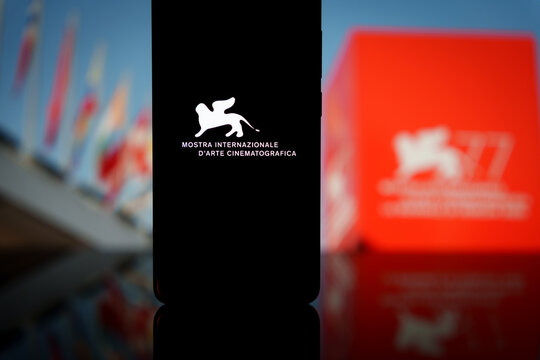 Venice Film Festival logo displayed on a smartphone screen with a blurred red festival banner and flags background, representing prestige cinema events and mobile film industry coverage