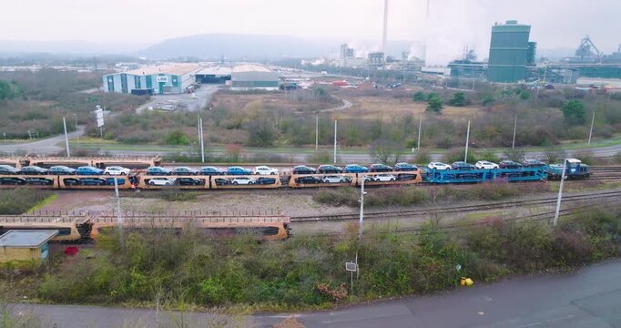 Wide aerial view of a long freight train transporting new cars on specialized auto rack wagons through an industrial area