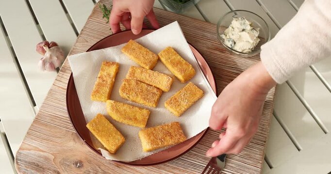 Crispy fried polenta sticks served on wooden board with creamy dip. Hands placing plate of golden cornmeal fries for rustic appetizer or side dish in bright kitchen setting.