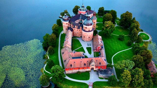 Gripsholm Castle, located in Mariefred, Sweden, is viewed from above. The castle features round towers with dark domes and reddish stone walls, surrounded by lush greenery. 