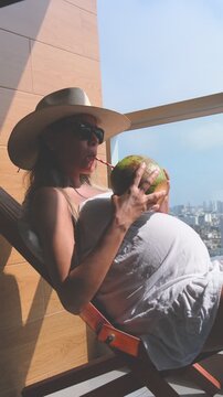 Expectant mother wearing a hat relaxing in a chair on a sunny balcony and drinking fresh coconut