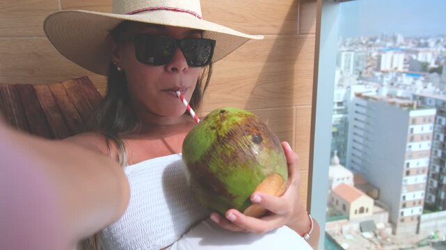 Woman taking a selfie while drinking coconut water from a straw on a balcony with a city view