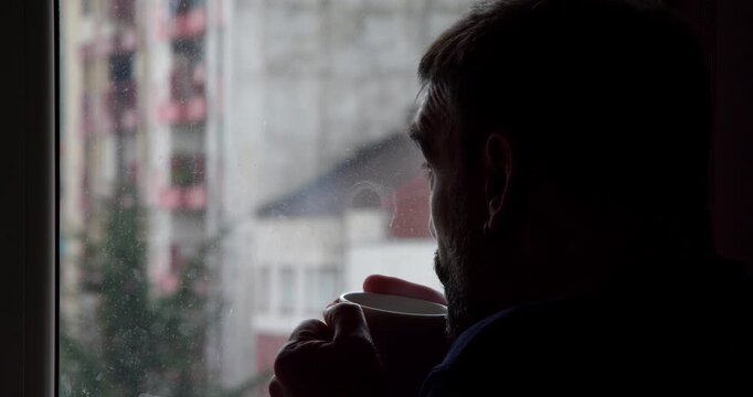 Solitary Contemplation by Window: A man gazes pensively out of a window, silhouetted against a backdrop of a blurred cityscape, lost in thought while holding a cup