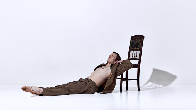 Male dancer lying on floor near chair in relaxed pose in minimal studio. Concept of creative campaign visuals, fashion storytelling, editorial content, and artistic lifestyle branding