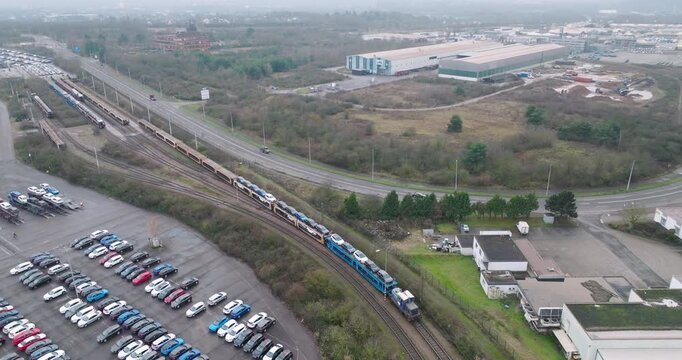 Wide aerial view of a long freight train transporting new cars on specialized auto rack wagons through an industrial area