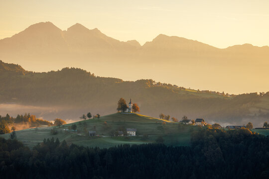 View of a hilltop church bathed in golden light amidst rolling hills and distant mountains, evoking tranquility and spiritual serenity, Selca, Slovenia.