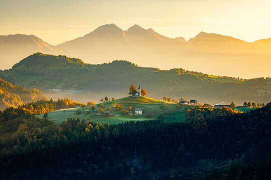 View of a hilltop church bathed in the golden light of dawn, overlooking rolling hills and distant mountains, Selca, Skofja Loka, Slovenia.