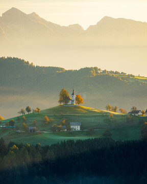 View of a church atop a verdant hill, surrounded by trees with hints of autumn colors, against a backdrop of misty mountains, Selca, Skofja Loka, Slovenia.