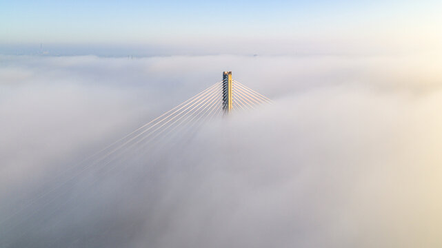 Aerial view of a bridge pierces the dense, ethereal fog, its cables like sunlit threads against the muted sky, Warsaw, Masovian Voivodeship, Poland.