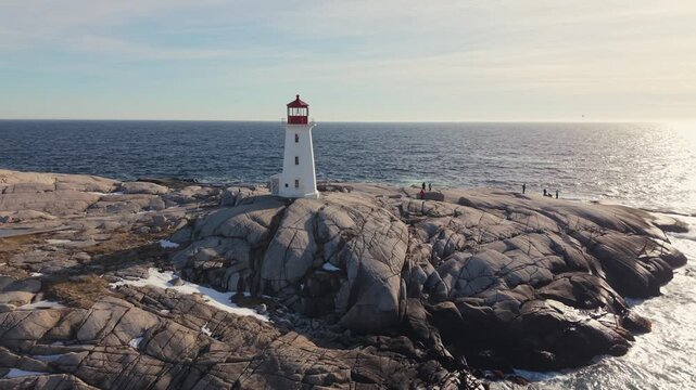 Drone aerial perspective: snow-covered boulders and Peggy's Cove lighthouse glow in warm sunset light. Atlantic coastline near Halifax, Nova Scotia Canada, peaceful winter evening scene.