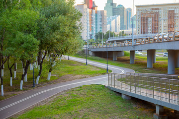 Fototapeta premium Bicycle path in city park winding under highway overpass. Pedestrian walkway near concrete flyover with metal railing. Urban landscape with road infrastructure and green trees in city.