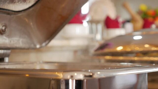 A female hand lifts a chafing dish lid releasing steam from hot prepared dishes arranged along a hotel breakfast serving counter indoors. Hotel breakfast.