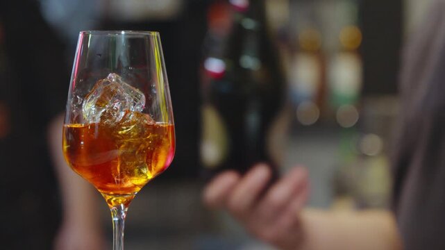 Bartender pouring sparkling wine into glass with ice and orange aperitif.