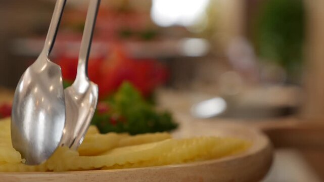 A guest uses serving tongs to place slices of cheese onto a plate from a breakfast buffet counter arranged with prepared dishes in a restaurant setting. Food selection.