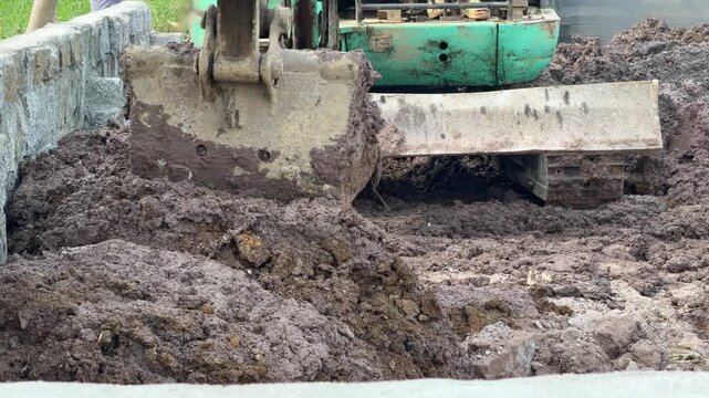 An excavator bucket moves loose soil across a construction site surface during ground shaping operations in an outdoor building preparation area. Earthmoving operations.