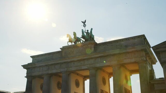 BERLIN, GERMANY - MARCH 23 2026: Panning shot close up of the Quadriga statue on top of the Brandenburg Gate with dramatic sunlight and lens flare in the background