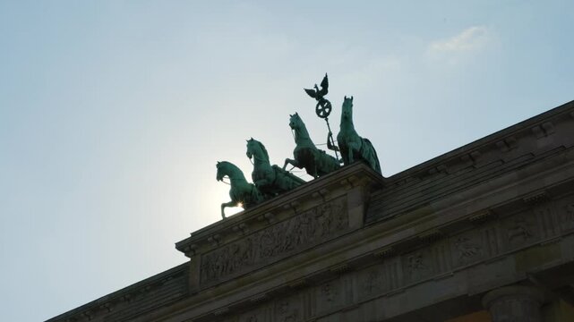 BERLIN, GERMANY - MARCH 23 2026: Panning shot close up of the Quadriga statue on top of the Brandenburg Gate with dramatic sunlight and lens flare in the background