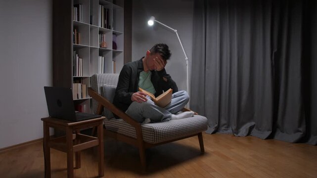 Overwhelmed young man sitting on a chair, holding a book, covering his face with his hand while feeling exhausted and stressed from prolonged studying or work in a dimly lit room at night