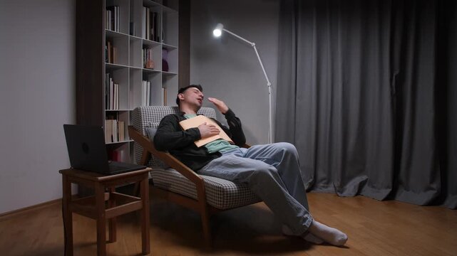 Young man sleeping in an armchair, holding a book on his chest, suffering from fatigue and exhaustion after late night studying under a lamp in his apartment