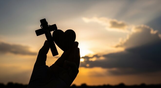 A hand holding a cross and a heart against a sunset sky.