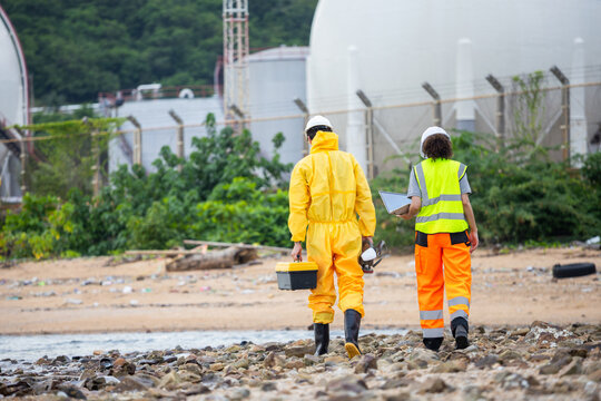 Environmental Response Team in Hazmat Suit and Safety Gear Walking on Polluted Shoreline, Technicians Carrying Equipment and Laptop for Chemical Spill Assessment Near Industrial Facility