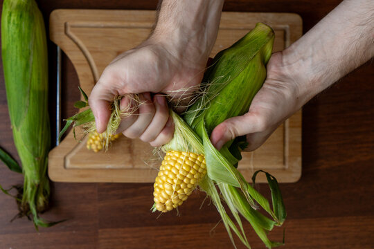Man's hands holding and shucking fresh corn on a wooden table. Top view