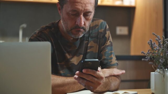 Mature adult caucasian male wearing camouflage army shirt using mobile phone at home, 4K