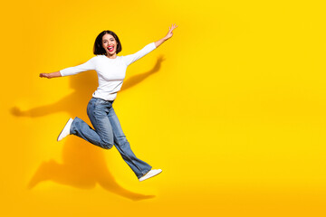 Obraz premium Happy young chic female jumping in white shirt and jeans against bright yellow background for lifestyle fashion advertising and promotion