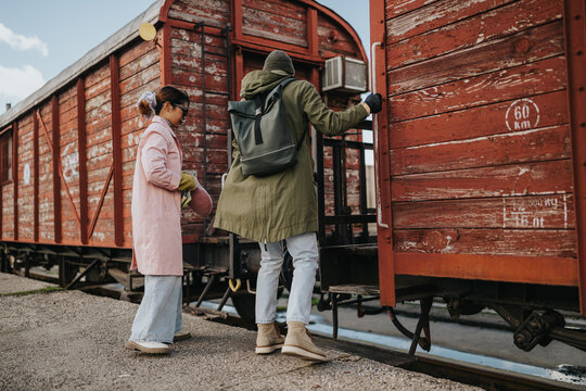 Two tourists board a weathered red freight train car at a small platform. A couple prepares to travel beside an old wooden boxcar in a quiet station.