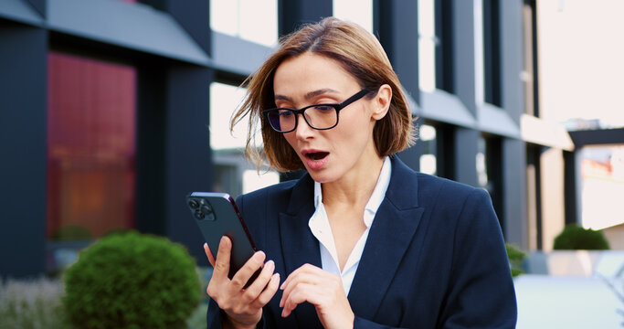 Frustrated professional businesswoman holding a smartphone, frowning and reacting to bad news, showing stress and disappointment while standing outdoors near a modern office building.