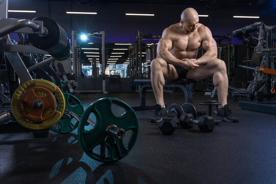 Muscular man rests on a weight bench surrounded by gym equipment, appearing contemplative after a workout.